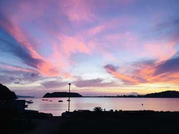 La imagen representa una serena puesta de sol sobre una masa de agua, con barcos y una farola, con una silueta de tierra en primer plano y un cielo rosa y morado.