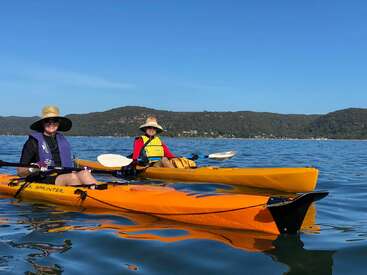 Dos personas en kayaks amarillos sobre una gran masa de agua, con sombreros y chalecos salvavidas, con un paisaje montañoso de fondo bajo un cielo azul despejado.