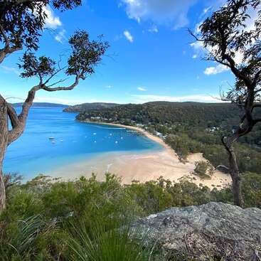 L'image représente une scène de plage sereine avec un rivage sablonneux, une eau bleue calme et une végétation luxuriante, sur fond pittoresque de collines et de ciel bleu clair.