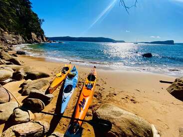 La imagen representa dos kayaks en una playa de arena, con una serena masa de agua y un cielo azul claro de fondo, evocando una sensación de tranquilidad.