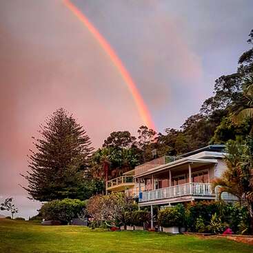 La imagen representa una serena escena de una casa con balcón, rodeada de frondosos árboles y un vibrante arco iris que se extiende por el cielo.