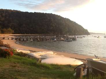 La imagen representa una serena escena playera con un muelle, barcas y exuberante vegetación, sobre el pintoresco fondo de una colina y un cielo nublado.