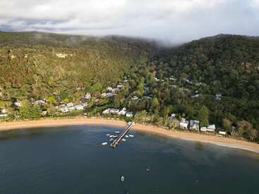 La imagen representa una serena ciudad costera enclavada entre exuberantes colinas verdes, con una playa de arena, un embarcadero y algunas barcas flotando en las tranquilas aguas.