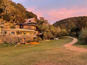 La imagen representa una serena casa de varios pisos con un gran patio y un sinuoso camino de tierra, enclavada entre árboles y colinas bajo un cielo rosa y azul.