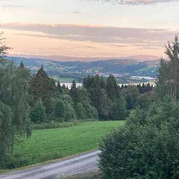Uma paisagem serena com campos verdes exuberantes, árvores frondosas, um lago ao longe, colinas onduladas e um céu colorido ao pôr do sol, com uma estrada rural sinuosa ao fundo.