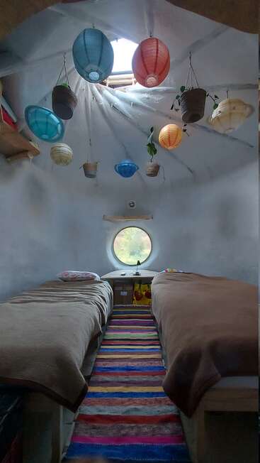 Cette chambre confortable comprend deux lits simples, des lanternes suspendues colorées, des plantes en pot, une fenêtre ronde, une lucarne, des étagères en bois et un tapis rayé aux couleurs vives. Atmosphère accueillante.