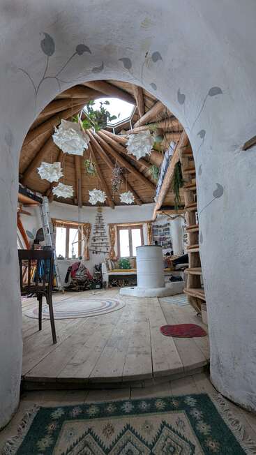 Une chambre rustique et confortable avec un plafond en bois en forme de dôme, des murs enduits de plâtre blanc, des plantes suspendues, une décoration artisanale, un escalier en colimaçon, de la lumière naturelle et un mobilier éclectique et chaleureux au charme authentique.