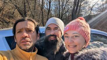 Trois personnes posent pour un selfie en plein air par une journée ensoleillée. Elles sont vêtues de vêtements d'hiver chauds et se tiennent devant une voiture, avec des arbres dénudés en arrière-plan.