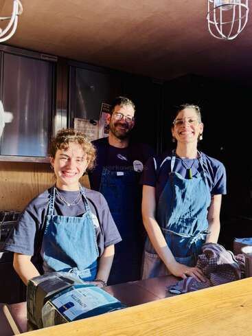 Trois personnes souriantes vêtues de tabliers bleus se tiennent derrière un comptoir en bois dans une cuisine, éclairées par la lumière chaude du soleil, semblant amicales et prêtes à aider les clients.
