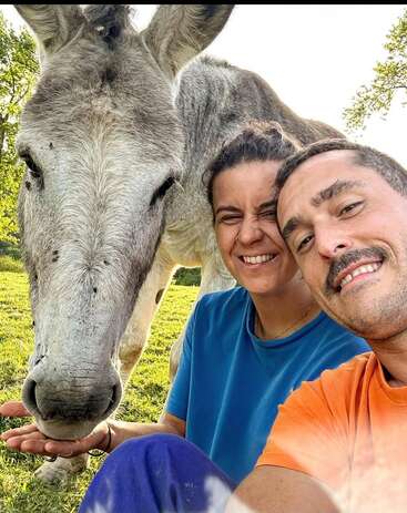 Um casal feliz tira uma selfie bem de perto ao ar livre com um burro simpático, todos sorrindo sob o sol forte, num cenário tranquilo e verdejante.