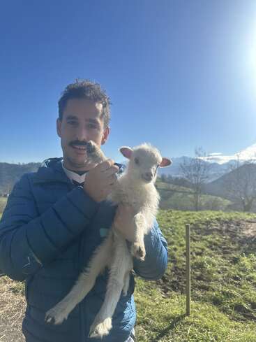 Um homem de jaqueta azul segura um pequeno cordeiro branco em um cenário ao ar livre ensolarado, com colinas, árvores e céu azul ao fundo.