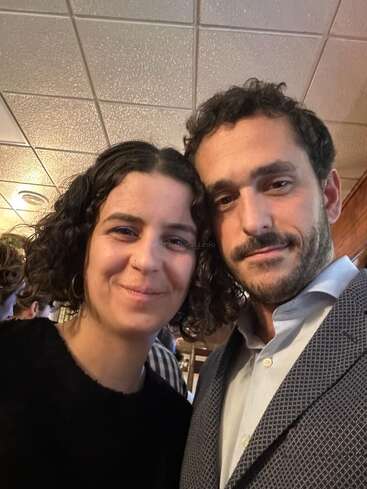 Um homem e uma mulher estão sorrindo bem pertinho um do outro para uma selfie em um ambiente interno com iluminação aconchegante; os dois parecem felizes e relaxados, vestidos com roupas casuais e formais.