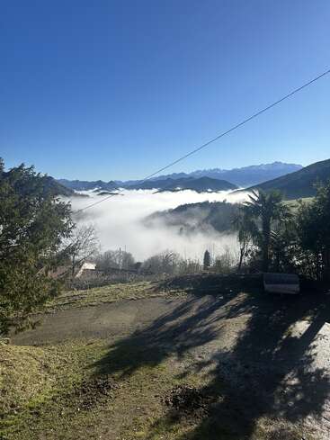 Un ciel bleu limpide surplombe une vallée embuée, avec des montagnes au loin. Des arbres encadrent un champ baigné de soleil, tandis qu'un banc et les ombres apportent de la profondeur et une atmosphère paisible.