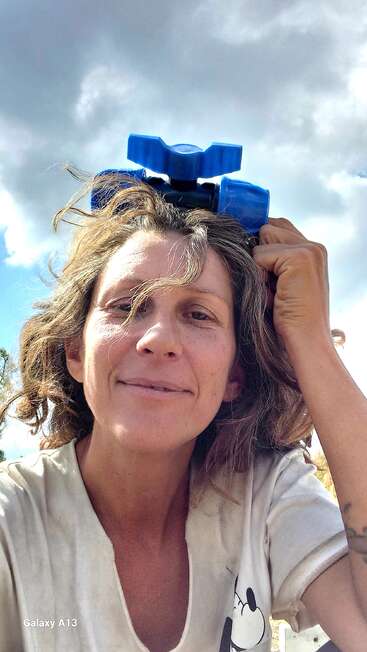 Une femme aux cheveux ébouriffés tient un jouet en plastique bleu sur sa tête, en souriant. Elle porte une chemise blanche à l'effigie de Mickey Mouse. Ciel nuageux et soleil en arrière-plan.