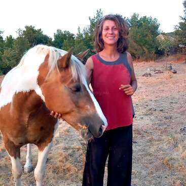 Una mujer sonriente se encuentra al aire libre sobre hierba seca junto a un caballo marrón y blanco, acariciándolo suavemente. Árboles y vegetación llenan el fondo, sugiriendo el campo.