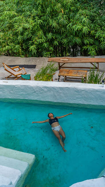 A woman floats peacefully in a clear blue pool. Bamboo plants surround the area. Nearby, wooden lounge chairs and tables create a relaxing, natural outdoor atmosphere.