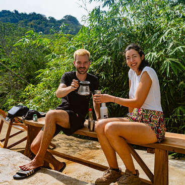 A man and woman sit on a wooden bench outdoors, smiling and sharing drinks, surrounded by lush green foliage and sunlight, enjoying a relaxed, cheerful moment together.