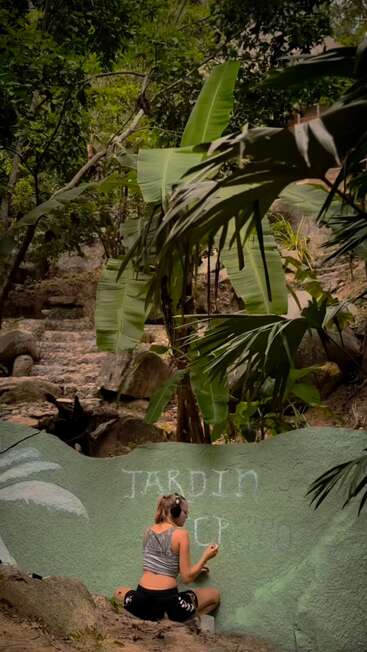 In a lush, green jungle setting, a woman wearing headphones is painting or writing "JARDIN" on a large green rock, surrounded by tropical plants.