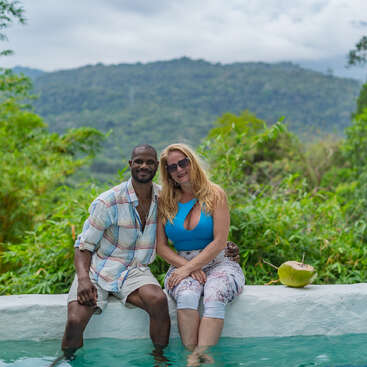 Una pareja sonriente está sentada al borde de una piscina, con los pies en el agua, rodeada de exuberante vegetación y montañas. Un coco descansa junto a ellos.