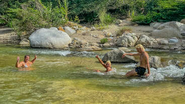 Three people enjoy swimming in a clear, shallow river surrounded by rocks and lush greenery. They smile and gesture peace signs, creating a playful, relaxed atmosphere.