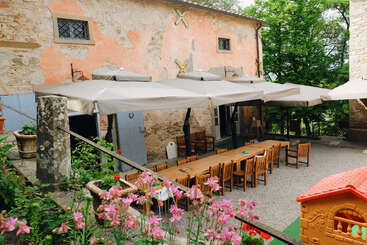 Cette image représente une charmante salle à manger en plein air avec des tables et des chaises en bois, de grands parasols, des fleurs épanouies, des murs en pierre rustiques et une petite maison de jeu en plastique pour les enfants.