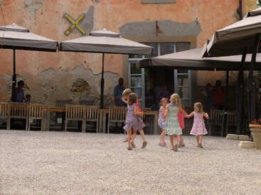 Cinq jeunes filles vêtues de robes colorées se tiennent par la main et jouent ensemble à l'extérieur, sous de grands parasols, sous le regard d'adultes installés derrière des tables en bois près d'un vieux bâtiment.