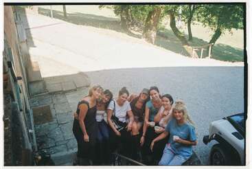 Un groupe de huit femmes est assis à l'extérieur, souriant à l'appareil photo. Certaines tiennent des tasses, profitant d'un moment de détente dans un cadre ensoleillé et verdoyant.