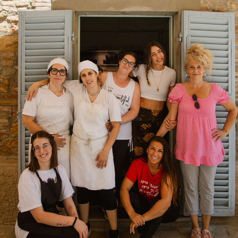 Sept femmes posent joyeusement devant une fenêtre aux volets bleus. Certaines portent des tabliers, d'autres des vêtements décontractés. Elles sourient, faisant preuve de camaraderie, de chaleur et d'une atmosphère détendue et joyeuse.