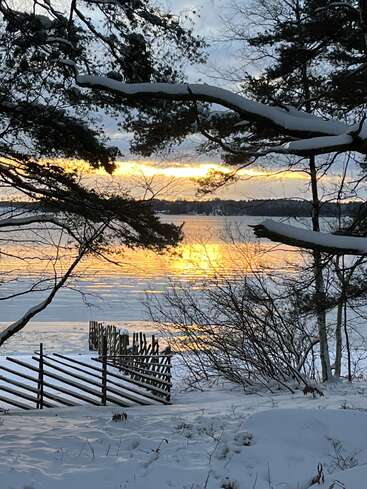 La neige recouvre le paysage et les arbres, tandis qu’un lac serein reflète un coucher de soleil doré. Une clôture rustique en bois et des branches dénudées encadrent cette paisible scène hivernale.