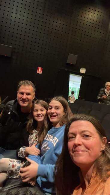 The image shows a family of four sitting in a dimly lit room with black walls and a speaker on the wall behind them, possibly at a movie theater.