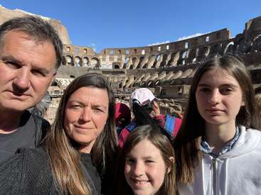The image shows a family of four posing for a photo in front of the Colosseum in Rome, Italy, with a clear blue sky above.
