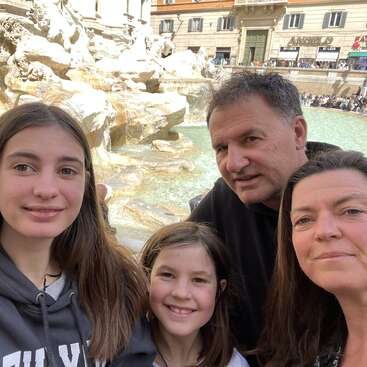 The image shows a family of four posing for a selfie in front of a large fountain with statues, likely in a European city.