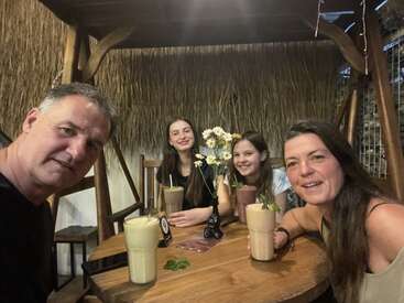 A family of four sits together at a wooden table in a cozy, rustic restaurant, enjoying milkshakes, smiling warmly, and posing for a selfie. Flowers decorate the table.