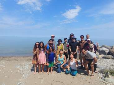 A diverse group of people, including children and adults, pose happily on a sandy beach near rocks and water, under a bright blue sky, some smiling.