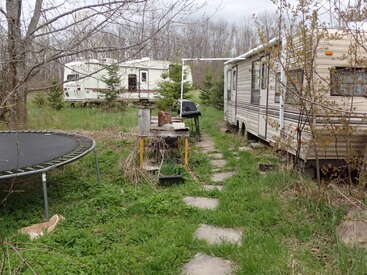 Two campers are parked in a grassy, wooded yard. Between them, there’s a stone path, a trampoline, an old table with clutter, and overgrown vegetation.