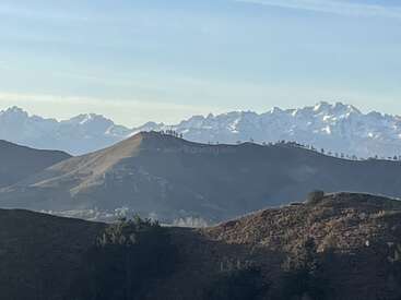 La imagen muestra unas colinas onduladas en primer plano, con árboles dispersos, y unas extensas montañas nevadas al fondo, bajo un cielo azul y despejado. Un paisaje tranquilo.