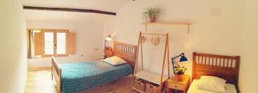 Este acogedor dormitorio cuenta con dos camas individuales de madera, una mesita de noche con una lámpara, una planta, una ventana con contraventanas y un perchero minimalista de madera.