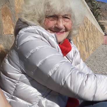 The image depicts an elderly woman with white hair, wearing a silver puffer jacket and red scarf, sitting on the ground in front of a stone wall.