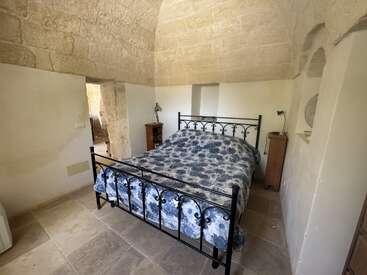 Dieses gemütliche Schlafzimmer mit Steinwänden verfügt über ein schmiedeeisernes Bett mit einer blauen Bettdecke mit Blumenmuster, zwei Nachttische aus Holz, eine Gewölbedecke und sanftes Tageslicht.