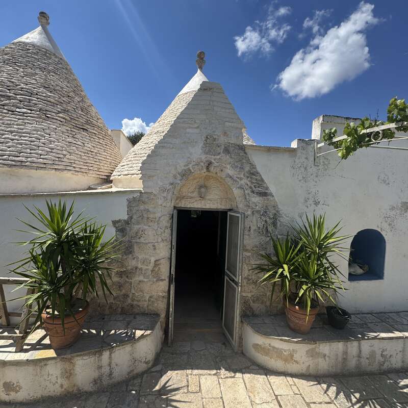 Dieses Bild zeigt ein traditionelles Trullo-Haus aus Stein mit kegelförmigen Dächern, strahlend blauem Himmel, Topfpflanzen am Eingang und sonnenbeschienenen Wänden, die eine rustikale, friedliche Atmosphäre schaffen.