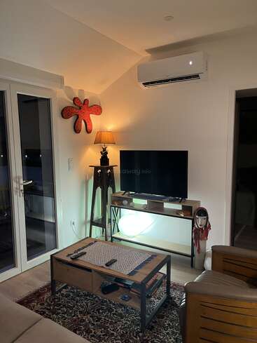 Uma sala de estar aconchegante com TV, mesa de madeira, poltrona, ar-condicionado, luminária, arte na parede, livros, máscara decorativa e um tapete estampado. Iluminação aconchegante.