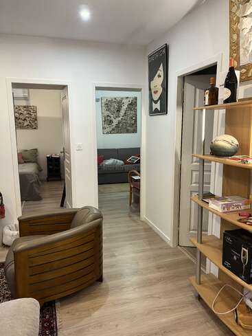 Um corredor aconchegante com piso de madeira, poltrona, prateleiras abertas com garrafas e decoração, três portas que levam aos quartos, arte moderna na parede e iluminação interna suave.