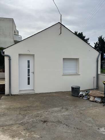 Esta imagem mostra um pequeno edifício branco com um telhado inclinado, uma porta fechada, uma janela fechada, lixo e detritos em uma superfície de concreto cinza.