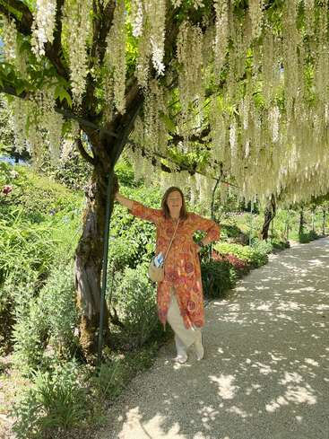 Uma mulher está em um caminho de jardim iluminado pelo sol, sob uma cascata de flores de glicínias brancas. Ela usa um vestido colorido e sorri, cercada por plantas verdes exuberantes.
