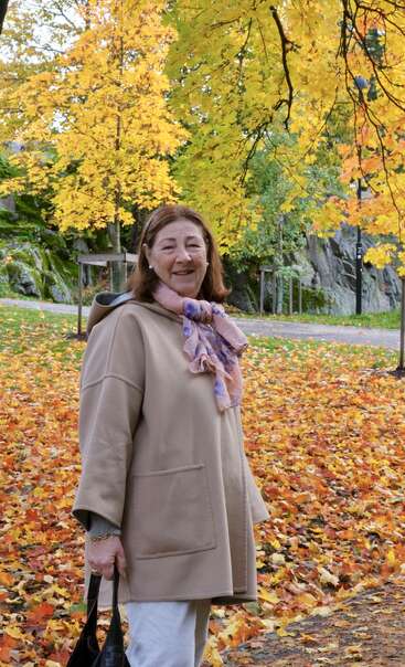 Uma mulher sorridente está em um parque repleto de folhas coloridas de outono. Ela usa um casaco bege, um cachecol rosa e segura uma bolsa preta, cercada por folhagens amarelas.