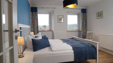Esta luminosa habitación cuenta con una cama blanca con detalles en azul, dos ventanas con vistas panorámicas, cortinas azules, lámparas de mesilla, cuadros y suelo de madera que le da un toque acogedor.