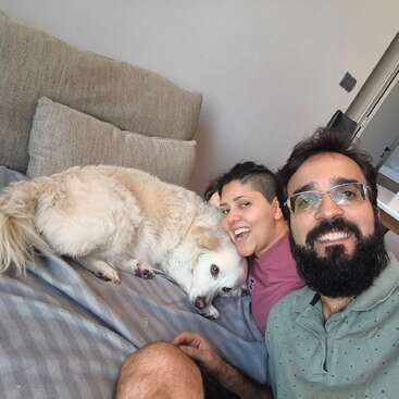 Ein fröhliches Paar posiert für ein Selfie mit seinem flauschigen weißen Hund, der auf dem Sofa liegt. Alle sehen glücklich und entspannt aus und genießen gemeinsam einen gemütlichen Moment zu Hause.