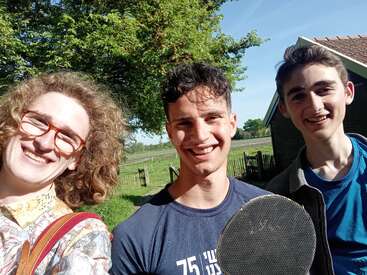 Drei lächelnde junge Menschen posieren an einem sonnigen Tag im Freien, mit Bäumen, einem Feld und einem Haus im Hintergrund. Einer hält einen Tischtennisschläger in der Hand.