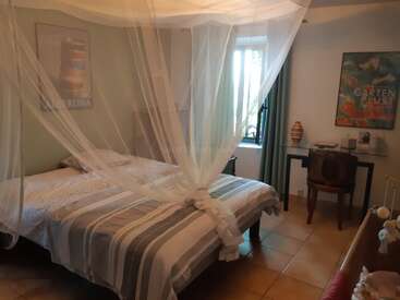 This cozy bedroom features a double bed with a mosquito net, striped bedding, a desk with a chair, wall art, window, and soft natural light.
