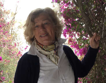A woman with blonde curly hair smiles, standing among vibrant pink flowers. She wears a white shirt, dark cardigan, and patterned scarf, exuding warmth and elegance outdoors.